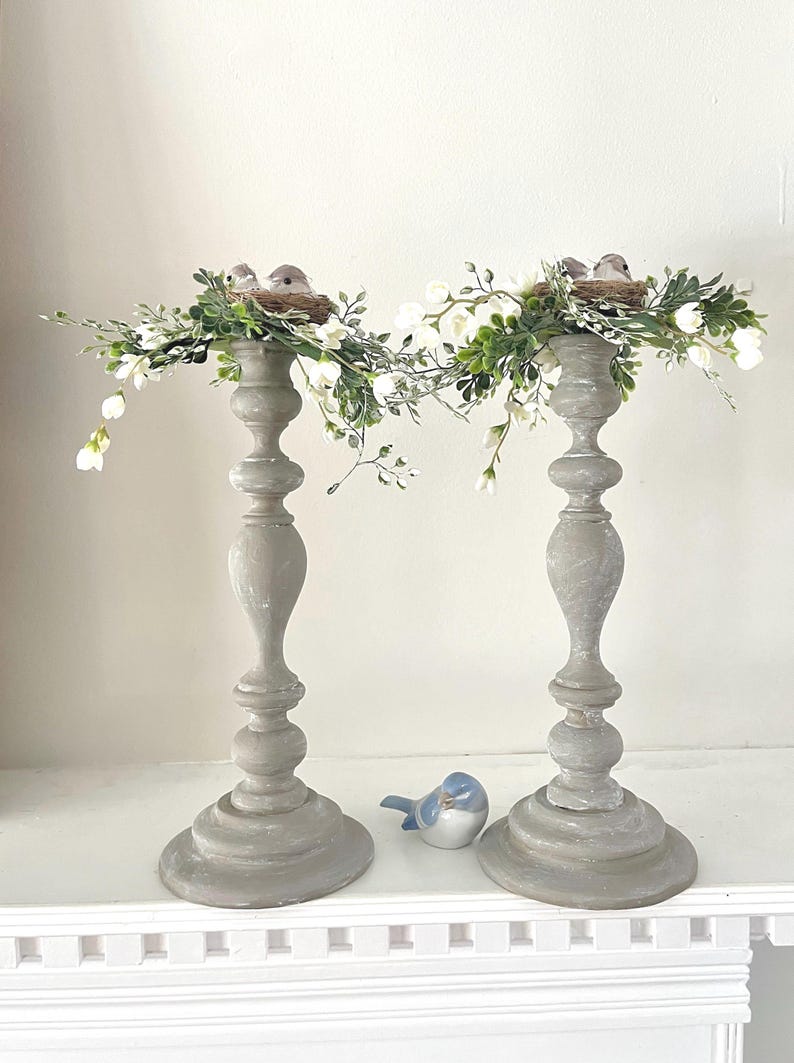 Pu&ograve; includere: Due portacandele in legno grigio con una ghirlanda floreale bianca e verde e piccoli nidi di uccelli in cima. I portacandele sono su uno scaffale bianco.