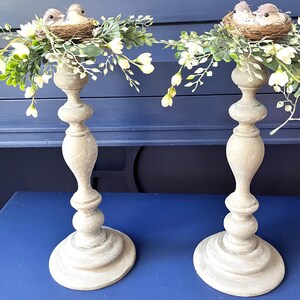 Pu&ograve; includere: Due portacandele in legno con una finitura bianca invecchiata. Ogni portacandele &egrave; sormontato da un nido di ramoscelli e due piccoli uccelli. I nidi sono circondati da una ghirlanda di fiori bianchi e foglie verdi.