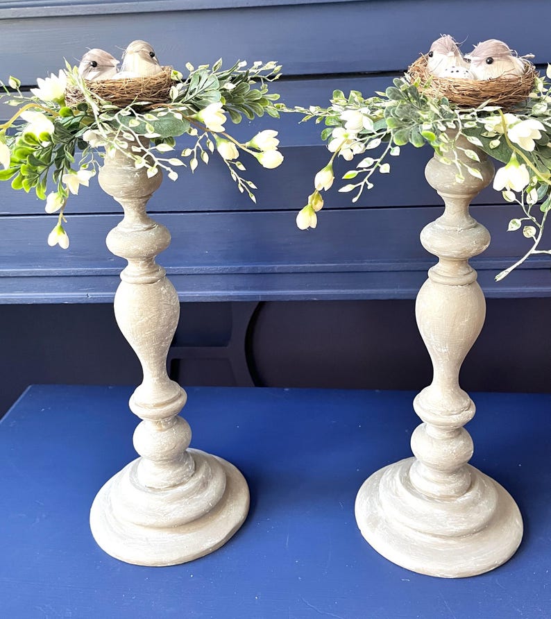 Pu&ograve; includere: Due portacandele in legno con una finitura bianca invecchiata. Ogni portacandele &egrave; sormontato da un nido di ramoscelli e due piccoli uccelli. I nidi sono circondati da una ghirlanda di fiori bianchi e foglie verdi.