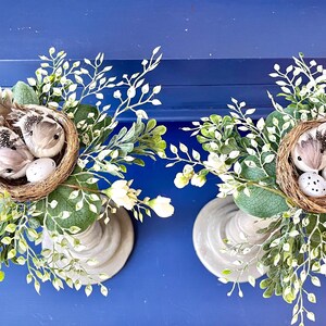 Pu&ograve; includere: Due portacandele grigi con nidi di uccelli bianchi finti e uccelli bianchi finti. Ogni nido &egrave; decorato con uova bianche finte. I portacandele sono circondati da verde finto verde e bianco.