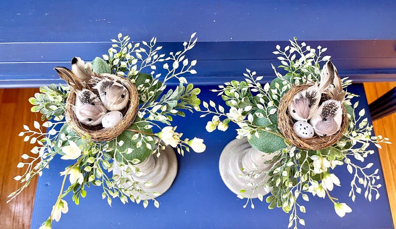 Pu&ograve; includere: Due portacandele grigi con nidi di uccelli bianchi finti e uccelli bianchi finti. Ogni nido &egrave; decorato con uova bianche finte. I portacandele sono circondati da verde finto verde e bianco.