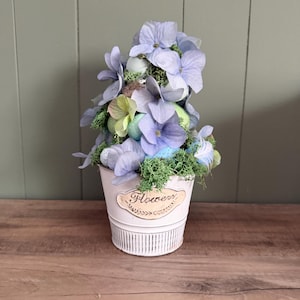 May include: A decorative arrangement of light purple hydrangea flowers and pastel-colored eggs in a white metal bucket. The bucket has a floral design and the word "Flower" on it. The arrangement sits on a wooden surface.