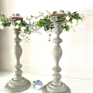Pu&ograve; includere: Un paio di portacandele in legno grigio con un nido di uccelli e uccelli in cima, decorati con foglie verdi e fiori bianchi.
