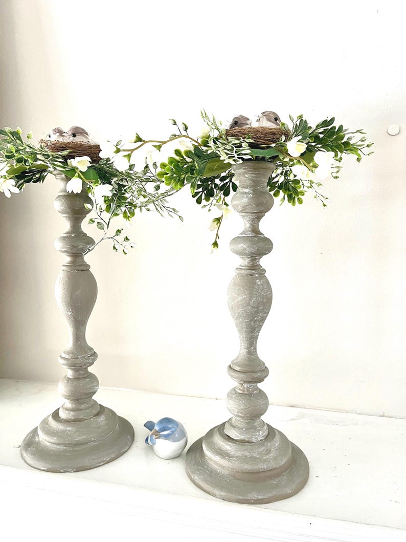 Pu&ograve; includere: Un paio di portacandele in legno grigio con un nido di uccelli e uccelli in cima, decorati con foglie verdi e fiori bianchi.