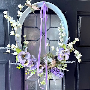 May include: Oval-shaped wreath with a light blue frame, adorned with purple pansies, white flowers, and a bird's nest with eggs. Purple ribbon hangs from the top. The wreath is displayed on a black door.