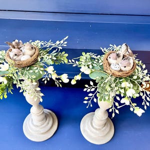 Pu&ograve; includere: Due portacandele in legno grigio con nidi di uccelli finti e fiori bianchi. Ogni nido contiene due piccoli uccelli e un uovo bianco.