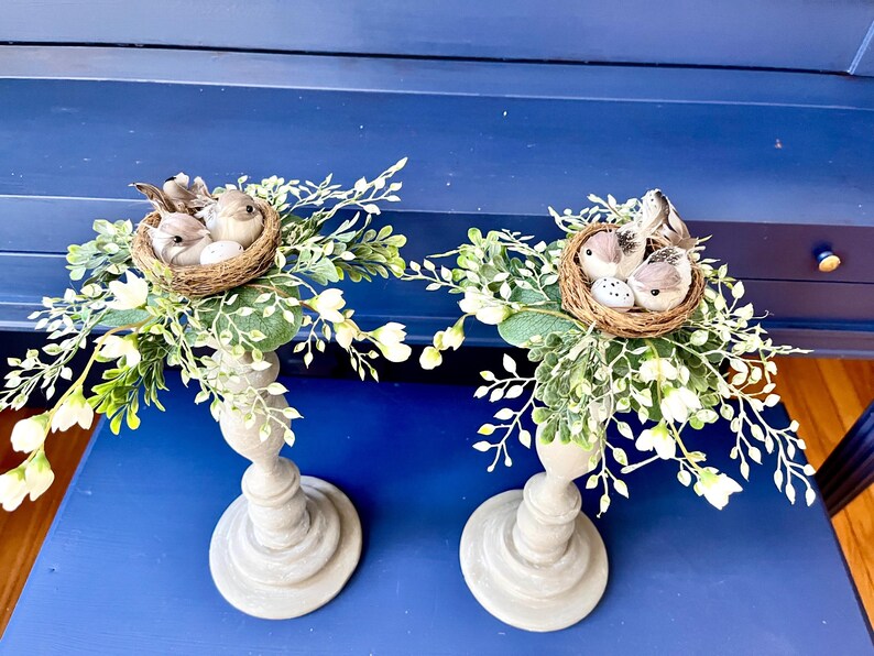 Pu&ograve; includere: Due portacandele in legno grigio con nidi di uccelli finti e fiori bianchi. Ogni nido contiene due piccoli uccelli e un uovo bianco.