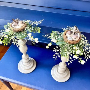 Pu&ograve; includere: Due portacandele in legno grigio con un nido di uccelli e del verde in cima. I nidi sono fatti di ramoscelli e contengono piccoli uccelli marroni e uova. Il verde &egrave; un mix di foglie verdi e fiori bianchi.