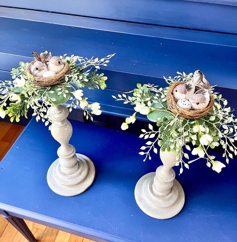 Pu&ograve; includere: Due portacandele in legno grigio con un nido di uccelli e del verde in cima. I nidi sono fatti di ramoscelli e contengono piccoli uccelli marroni e uova. Il verde &egrave; un mix di foglie verdi e fiori bianchi.