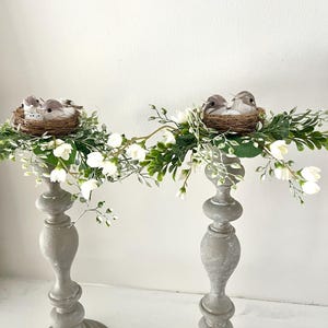 Pu&ograve; includere: Due portacandele in legno grigi con fiori bianchi e nidi di uccelli con uccelli in cima.