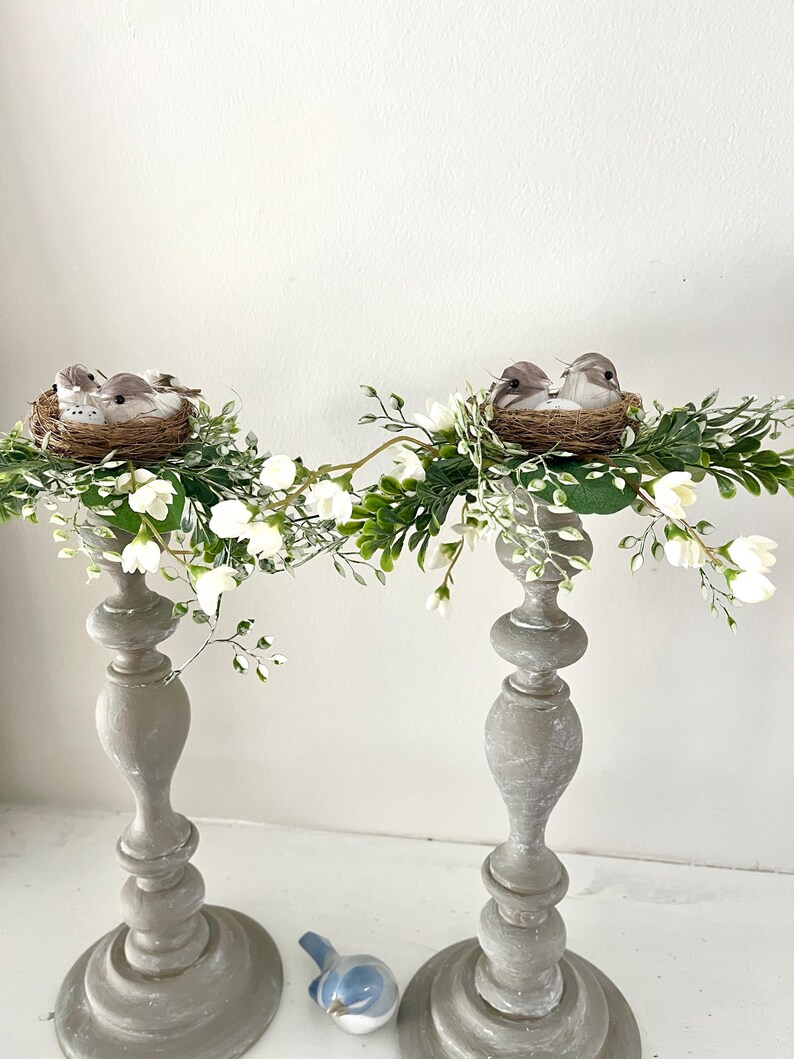 Pu&ograve; includere: Due portacandele in legno grigi con fiori bianchi e nidi di uccelli con uccelli in cima.