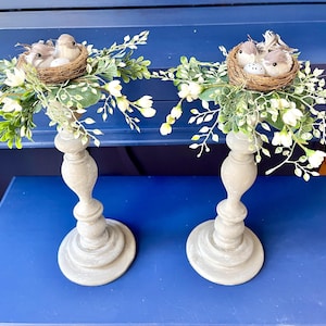 Pu&ograve; includere: Due portacandele grigi con fiori finti bianchi e nidi di uccelli con piccoli uccelli. I portacandele sono su una superficie blu.