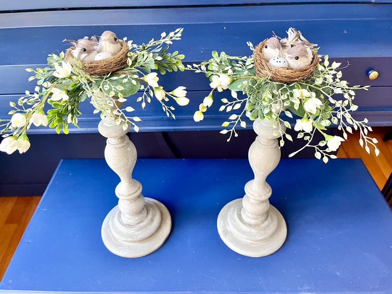 Pu&ograve; includere: Due portacandele grigi con fiori finti bianchi e nidi di uccelli con piccoli uccelli. I portacandele sono su una superficie blu.