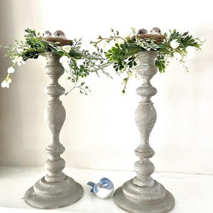 Pu&ograve; includere: Due portacandele in legno grigio con un nido di uccelli e del verde in cima. Il verde &egrave; una ghirlanda di piccoli fiori bianchi e foglie verdi. Una statuetta di un uccello blu &egrave; in primo piano.