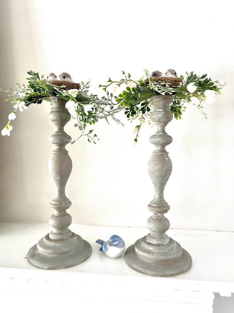 Pu&ograve; includere: Due portacandele in legno grigio con un nido di uccelli e del verde in cima. Il verde &egrave; una ghirlanda di piccoli fiori bianchi e foglie verdi. Una statuetta di un uccello blu &egrave; in primo piano.