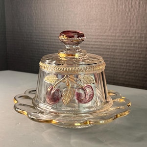 May include: A clear glass butter dish with a domed lid featuring a gold-toned rope trim and a purple cherry motif with gold leaves. The lid has a purple knob handle, and the base has a scalloped edge with a gold-toned rim, creating a decorative piece.