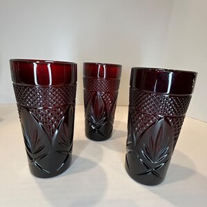 May include: Three ruby red drinking glasses. Each glass has a diamond-cut pattern on the upper part and a leaf design on the lower part. The glasses are tall and cylindrical, with a slightly flared rim.