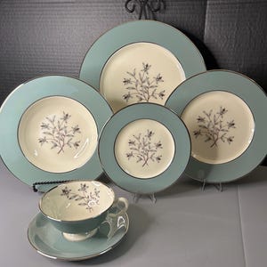 Könnte beinhalten: Ein Vintage-Geschirrset in Türkis und Creme. Das Set umfasst Teller in verschiedenen Größen, eine Teetasse und eine Untertasse. Jedes Stück hat ein Blumendesign in der Mitte und einen silbernen Rand.