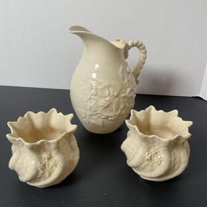 May include: A cream-colored ceramic pitcher with a floral design and a twisted handle, flanked by two matching small decorative pots. The pots and pitcher have a ruffled edge and embossed floral details. The set is displayed on a dark surface.