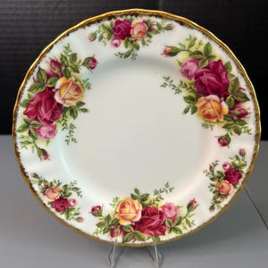 以下が含まれることがあります： 金色の縁とバラの花柄が施された白い磁器の皿。バラは赤、ピンク、黄色の色合いで、緑の葉が付いています。このお皿は装飾品です。