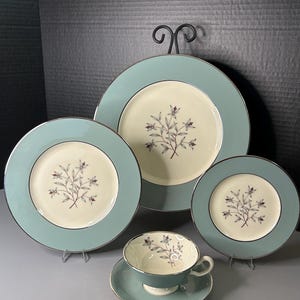 Könnte beinhalten: Ein Vintage-Geschirrset mit hellblauem Rand und cremefarbenem Mittelteil, verziert mit einem floralen Muster. Das Set umfasst Teller in verschiedenen Größen, eine Teetasse und eine Untertasse, alle mit silberner Zierleiste.