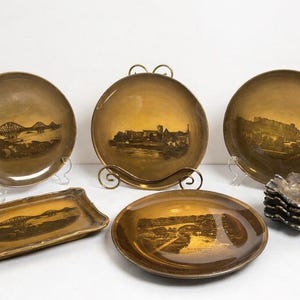 May include: A collection of vintage decorative plates and dishes with sepia-toned landscape scenes. Includes round plates, a rectangular tray, and small, scalloped dishes. The plates feature images of bridges and cityscapes.