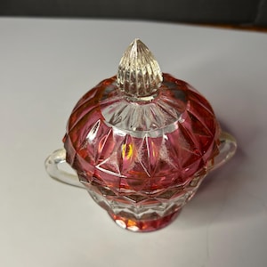 May include: A clear glass sugar bowl with a ruby red hue, featuring a faceted design. The bowl has a matching lid with a pointed finial and two small handles. The sugar bowl is a decorative kitchen item.