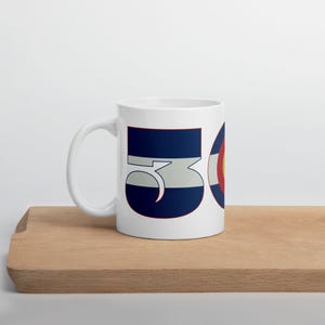 May include: White ceramic mug featuring the number "53" in navy blue with a light blue stripe, and a circular design with red, yellow, and white segments. The mug is on a wooden surface.