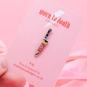 May include: A silver enamel pin in the shape of a knife with a black handle and a red rose design on the blade. The pin is displayed on a pink card with the text "worn to death apparel".
