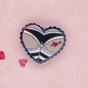 May include: Heart-shaped enamel pin featuring a stylized design of a rear view in black and tan underwear. A red heart with an arrow is tattooed on the rear. The pin has a silver scalloped border and is set against a pink background with red heart confetti.