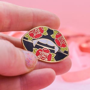 May include: A black and gold enamel pin in the shape of lips with fangs, featuring red roses and green leaves. The pin is held in a hand against a pink background. The lips have white fangs.