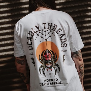Op de afbeelding: Wit T-shirt met een rugafbeelding met een spinnendesign, een oranje cirkel en de woorden "DEADLY THREADS" in een cirkelvormige boog. Onder de spin staat de tekst "WORN TO DEATH APPAREL".