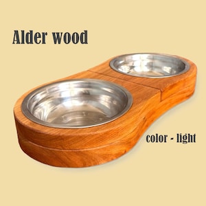 Könnte beinhalten: Ein doppelter Futternapf aus hellfarbenem Erlenholz. Der Holzsockel hält zwei Edelstahlnäpfe. Der Text "Alder wood" und "color - light" sind auf dem Bild zu sehen.