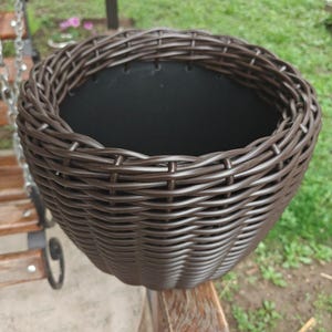 Puede incluir: Una cesta de plantación tejida marrón con un interior oscuro. La cesta tiene forma redonda y un diseño tejido texturizado. La jardinera está vacía y lista para plantas.