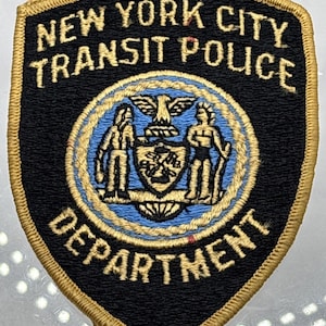 May include: A black and gold embroidered patch with the text "NEW YORK CITY TRANSIT POLICE DEPARTMENT". The patch features a blue circle with a crest and two figures. The patch has a gold border.