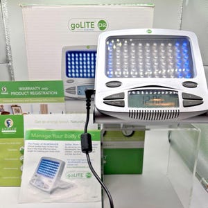 May include: A silver goLITE P2 device with a rectangular screen displaying blue and white lights. The device has buttons for light, timer, and power. The time 12:05 is displayed. The box and paperwork are visible in the background.