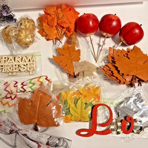 May include: A collection of fall-themed craft supplies, including orange and yellow artificial leaves, red apples on sticks, and decorative pumpkins. The image also shows the words "FARM FRESH" and "Love" in wooden cutouts.