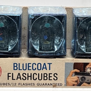 May include: A vintage package of Bluecoat Flashcubes, containing three flash cubes. Each cube is encased in a clear plastic shell with a blue tint. The packaging features the GE logo and an image of a woman taking a photo.