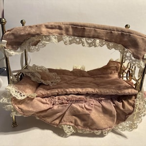 May include: A miniature bed with a canopy, featuring a gold-colored metal frame and a pink floral patterned fabric. The bed is adorned with white lace trim and bows, creating a delicate and ornate appearance. The bed's dimensions are approximately 12 inches long, 8 inches wide, and 10 inches high.