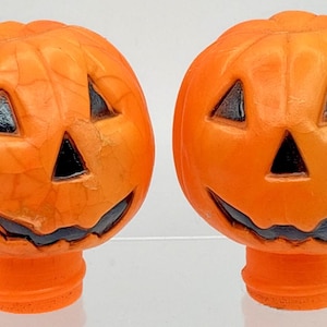 May include: Two orange pumpkin-shaped objects with carved facial features including triangular eyes, a triangular nose, and a wide, jagged mouth. The surfaces have a textured, cracked appearance, and each pumpkin sits atop a cylindrical base.