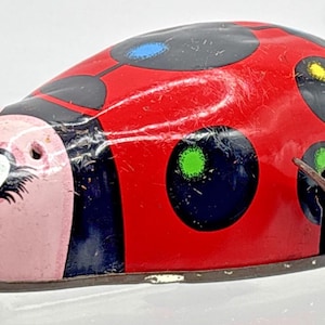 May include: A vintage-style tin wind-up toy shaped like a ladybug. The body is bright red with large black spots, some featuring small blue, yellow, and green centers. It has a light pink face with blue eyes and a metal winding key on the side.