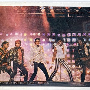 May include: A vintage photograph featuring five performers on a brightly lit stage with colorful circular lights. The central figure wears a white sequined jacket. The text at the bottom reads: PHOTO BY HARRISON FUNK © 1984 JEC.