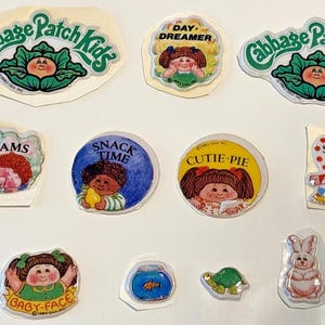May include: A collection of vintage Cabbage Patch Kids stickers. The stickers feature various characters and phrases like "Sweet Dreams," "Snack Time," and "Cutie-Pie." The color palette includes pastels, with images of babies and animals.