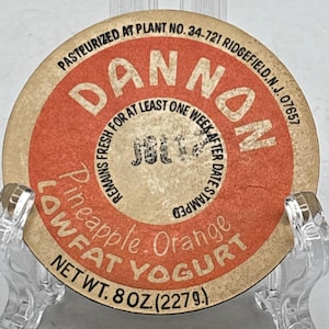 May include: A vintage Dannon yogurt lid with a red and tan color scheme. The lid features the Dannon logo, the words "Pineapple Orange Lowfat Yogurt," and the net weight of 8 oz (227g). The text "Pasteurized at Plant No. 34-721 Ridgefield, N.J. 07657" is also visible.