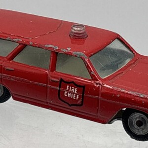 May include: A red die-cast toy station wagon fire chief vehicle with silver bumpers and wheels. The car features a small clear light on the roof and a black shield emblem on the side door with the white text FIRE CHIEF printed inside the shield.