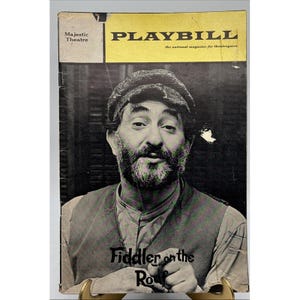 May include: A vintage Playbill magazine cover featuring a black and white portrait of a man with a beard and cap. The magazine has a yellow banner with the title "PLAYBILL" and the words "Fiddler on the Roof" at the bottom.