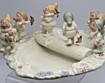 May include: A ceramic decorative tray featuring six small gnome figurines in cream-colored clothing with gold accents. The gnomes carry miniature pickaxes and a lantern, positioned around a central sculpted log on a base decorated with small flowers.