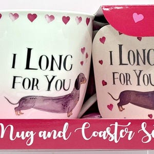 May include: A white mug and coaster set with a dachshund illustration and the text "I Long For You." The mug and coaster are decorated with pink hearts. The set is packaged in a red box.