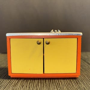 May include: A miniature orange and yellow cabinet with two doors and brass-colored knobs. The cabinet has a silver-colored countertop. The cabinet is a toy or decorative item.