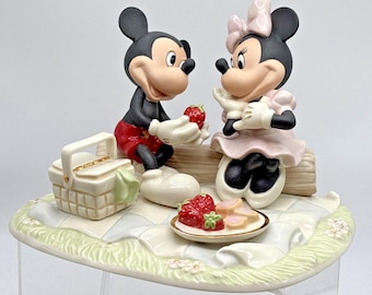 May include: A ceramic figurine featuring Mickey and Minnie Mouse sitting on a log during a picnic. Mickey holds a red strawberry, while Minnie sits beside him. A picnic basket and a plate with strawberries and cookies sit on a checkered blanket.